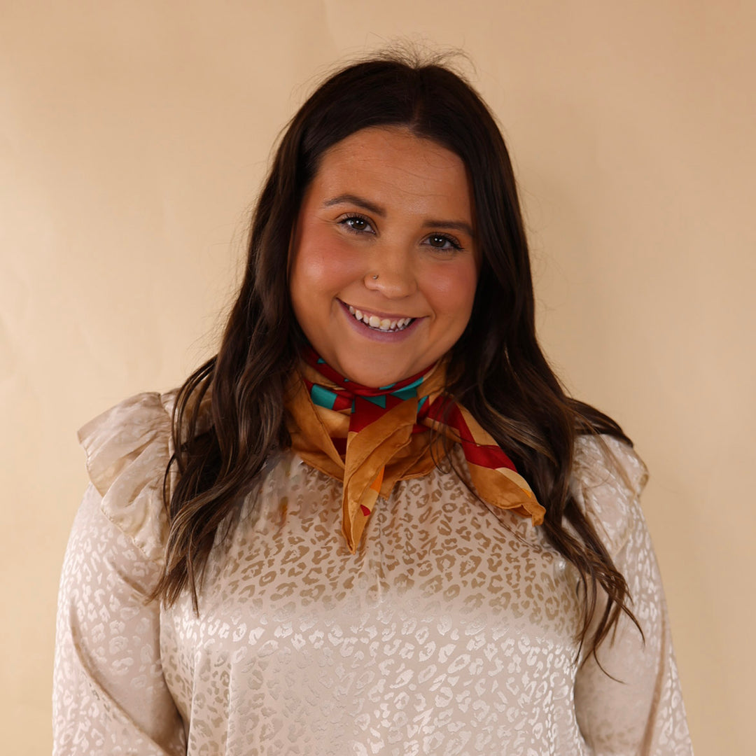 Brunette model wearing an ivory, leopard print top with gold and red design scarf tied around her neck. Model is pictured in front of a beige background. 