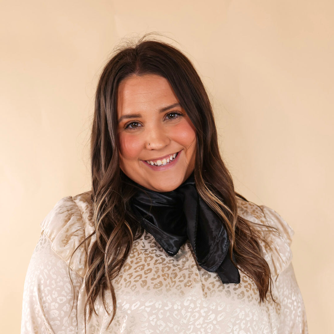 Brunette model wearing a ivory, leoaprd print top with a black scarf tied around her neck. Model is pictured in front of a beige background. 