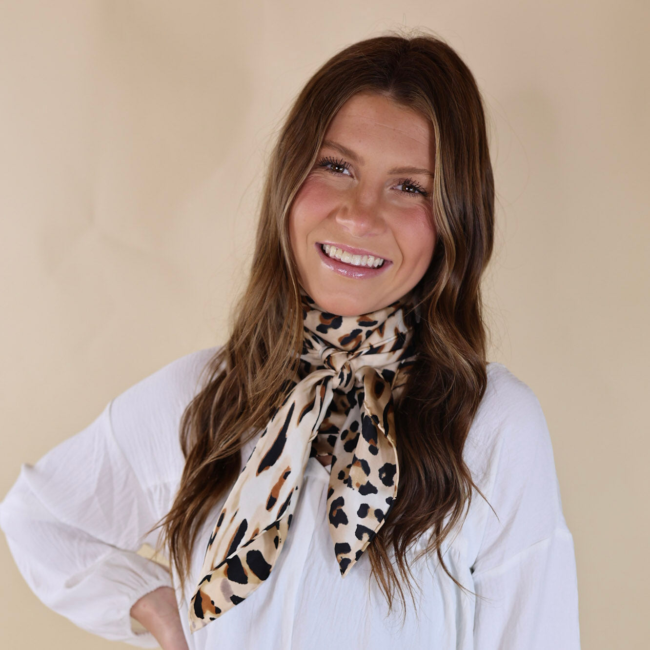 Brunette model wearing a white, off the shoulder top with a beige scarf with a brown and blakc leopard print tied around her neck. Model is pictured in front of a beige background. 
