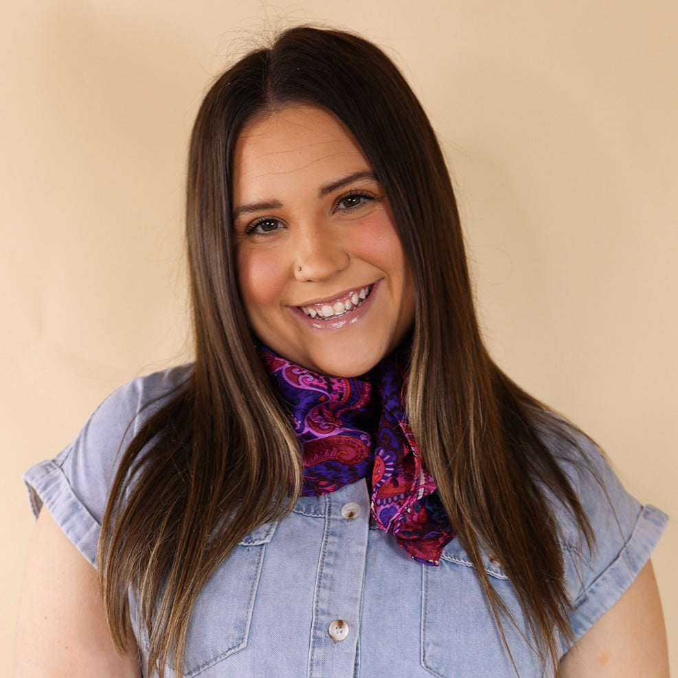 Brunette model is pictured wearing a denim button up top and a purple and pink mix paisley printed scarf tied around her neck. She is pictured in front of a beige background. 