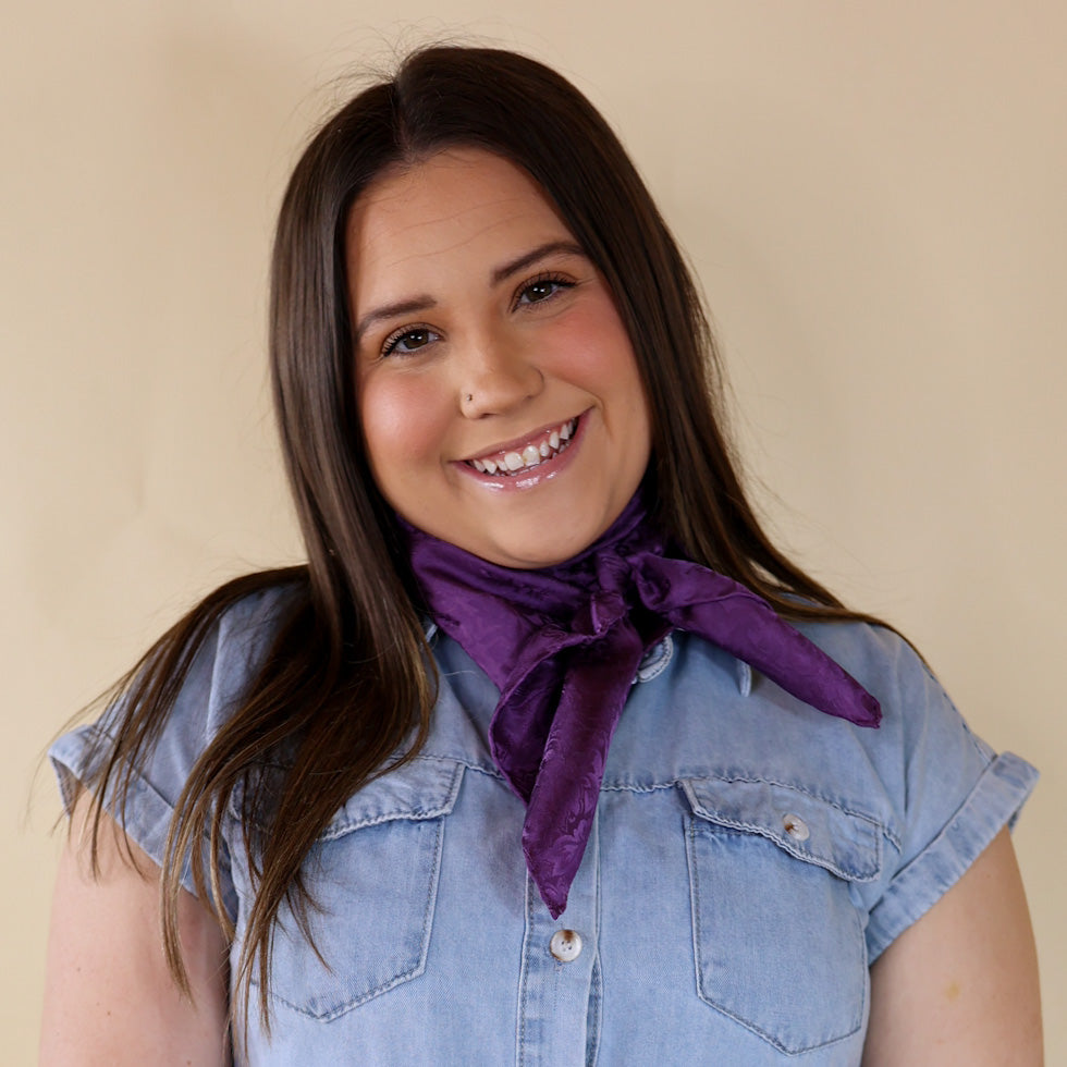 Brunette model is pictured wearing a denim button up top and a purple and jacquard printed scarf tied around her neck. She is pictured in front of a beige background. 