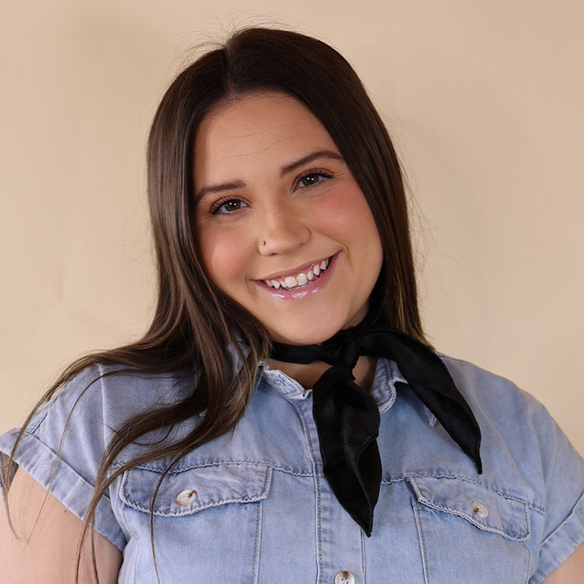 Brunette model wearing a short sleeve, denim button up with a black jacquard print scarf tied around her neck. This model is pictured in front of a beige background. 