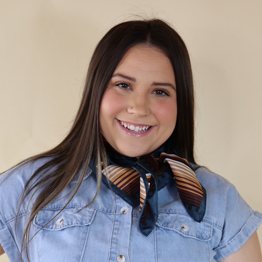 Brunette model is pictured wearing a denim button up top and a blue and brown striped scarf tied around her neck. She is pictured in front of a beige background. 