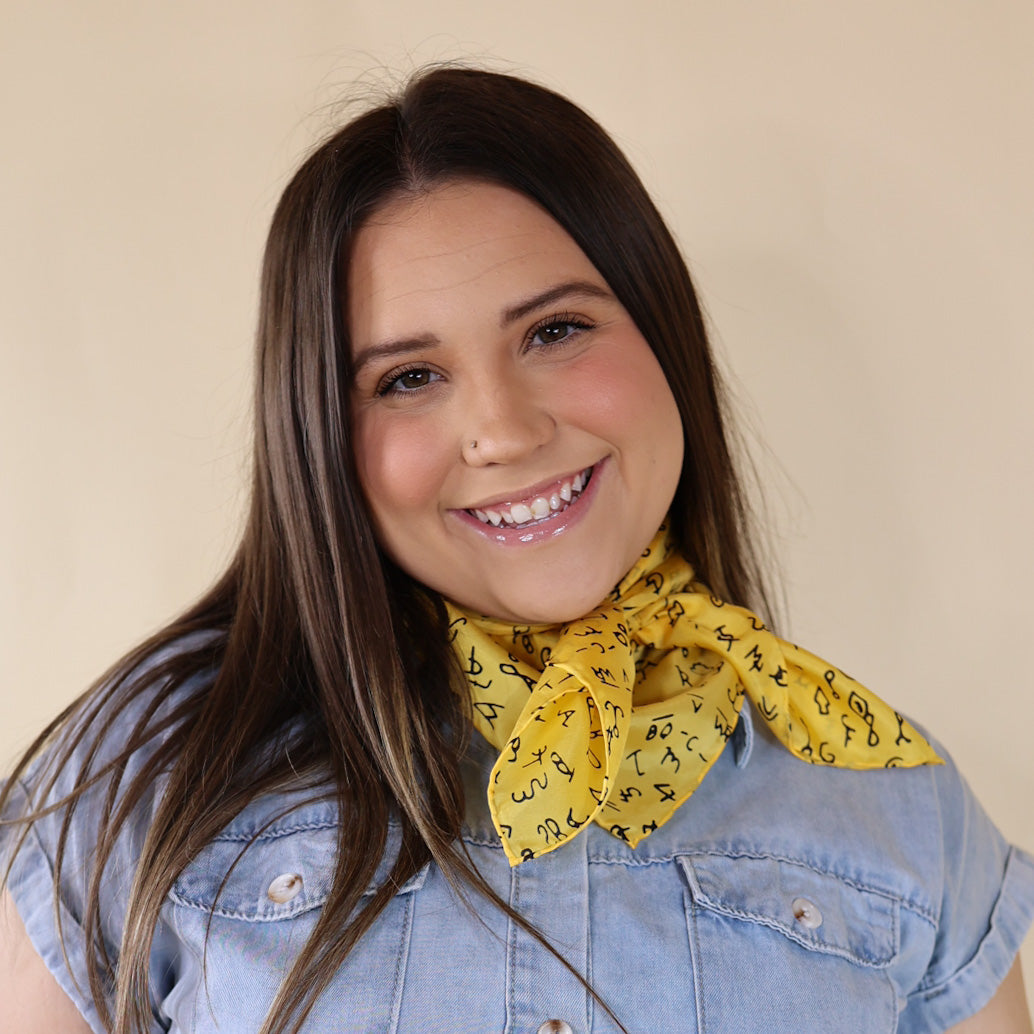 Brunette model is pictured wearing a denim button up top and a yellow with black symbols printed scarf tied around her neck. She is pictured in front of a beige background. 