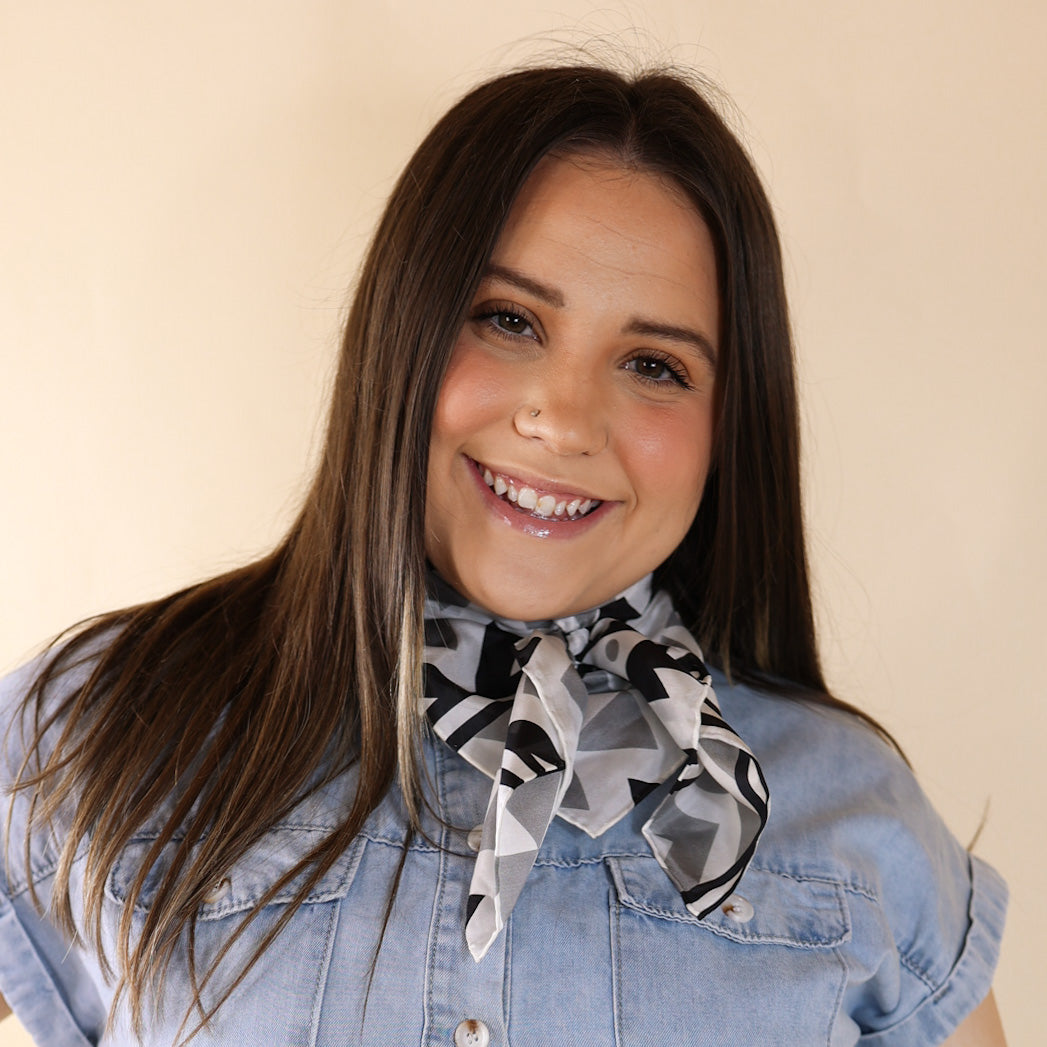 Brunette model wearing a short sleeve, denim button up with a black, white, and gey aztec print scarf tied around her neck. This model is pictured in front of a beige background. 