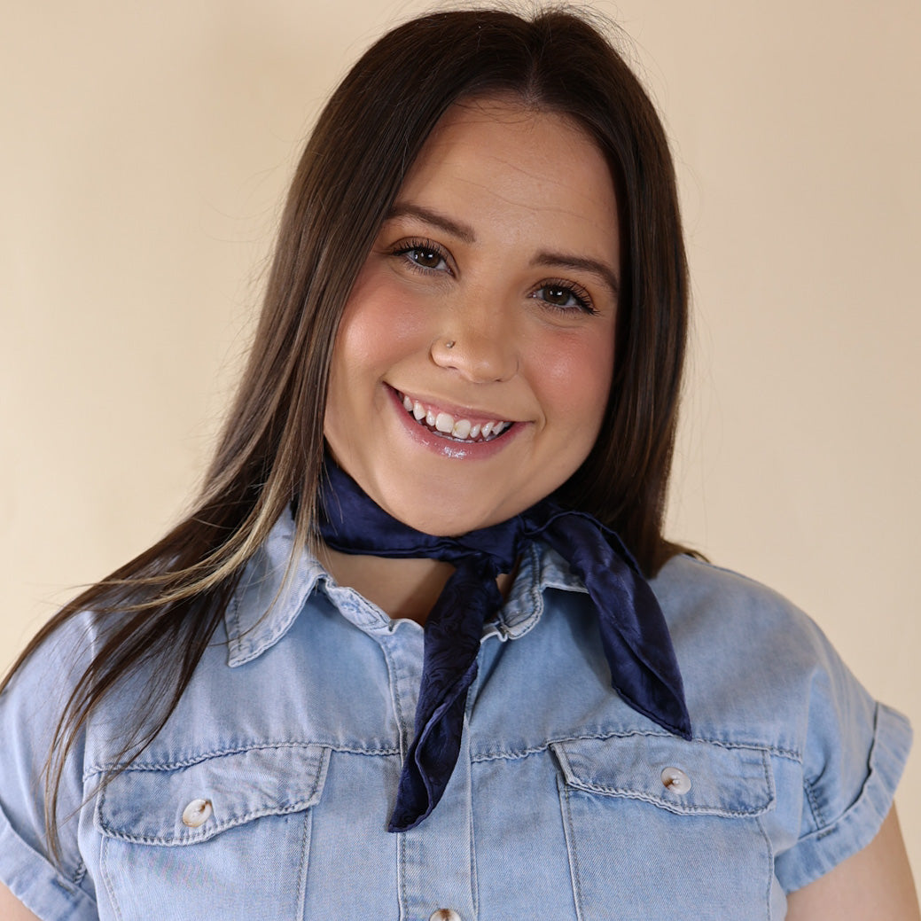 Brunette model wearing a short sleeve, denim button up with a navy blue jacquard print scarf tied around her neck. This model is pictured in front of a beige background. 