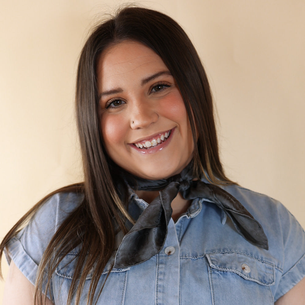 Brunette model is pictured wearing a denim button up top and a charcoal grey, jacquard printed scarf tied around her neck. She is pictured in front of a beige background. 