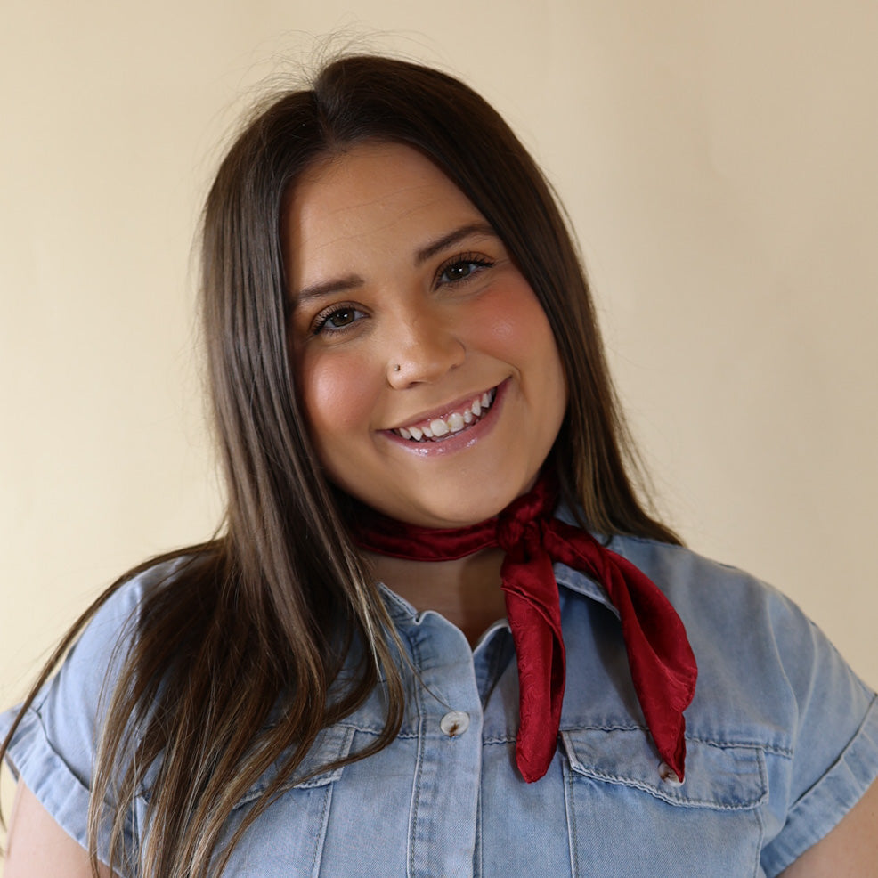 Brunette model is pictured wearing a denim button up top and a red, jacquard printed scarf tied around her neck. She is pictured in front of a beige background. 