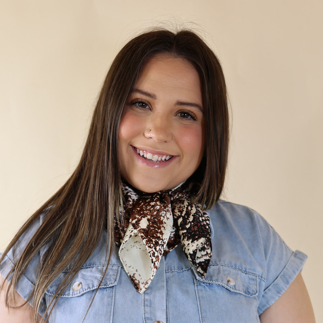 Brunette model wearing a blue dress with Brown and White animal print scarf tied around her neck. Model is pictured in front of a beige background.