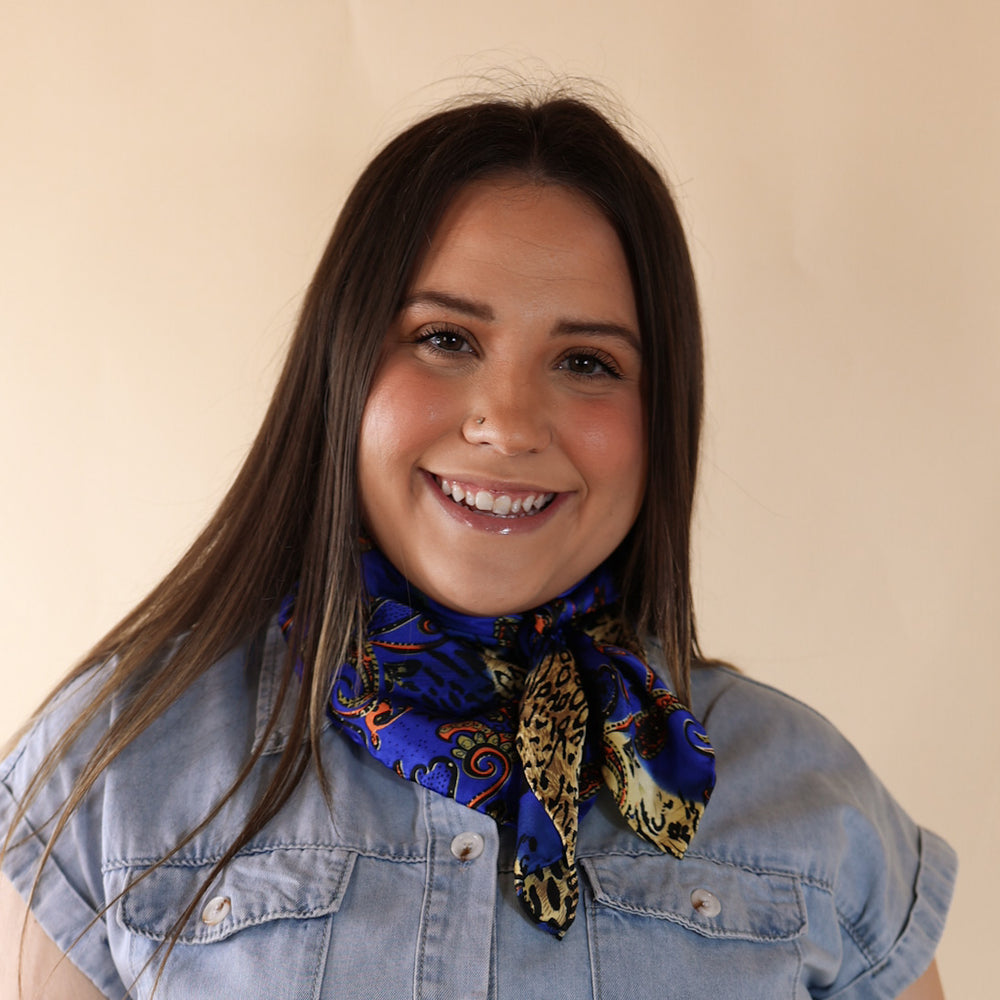 Brunette model wearing a blue dress with Royal Blue peacock print scarf tied around her neck. Model is pictured in front of a beige background.