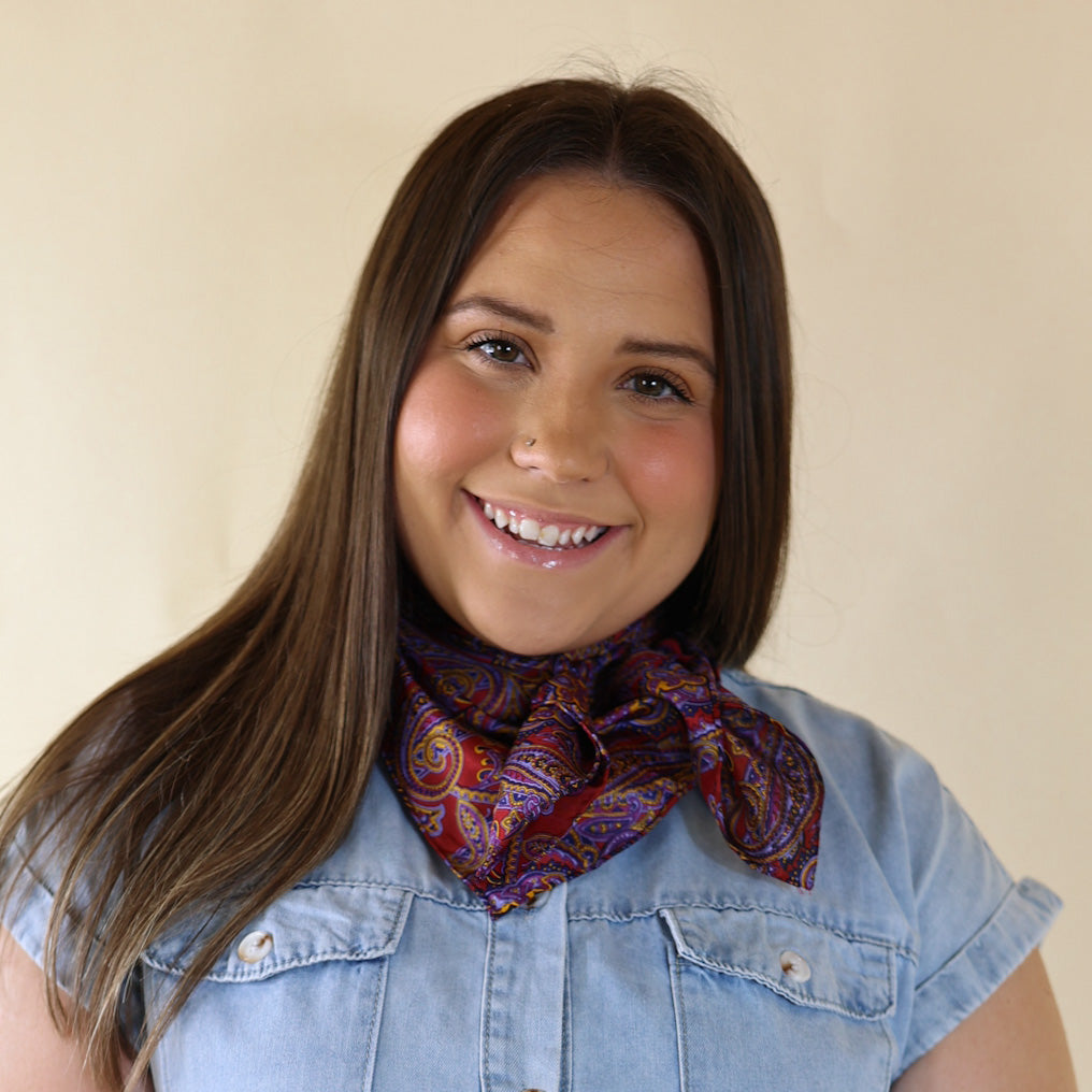 Brunette model is pictured wearing a denim button up top and a wine and navy mix paisley printed scarf tied around her neck. She is pictured in front of a beige background. 