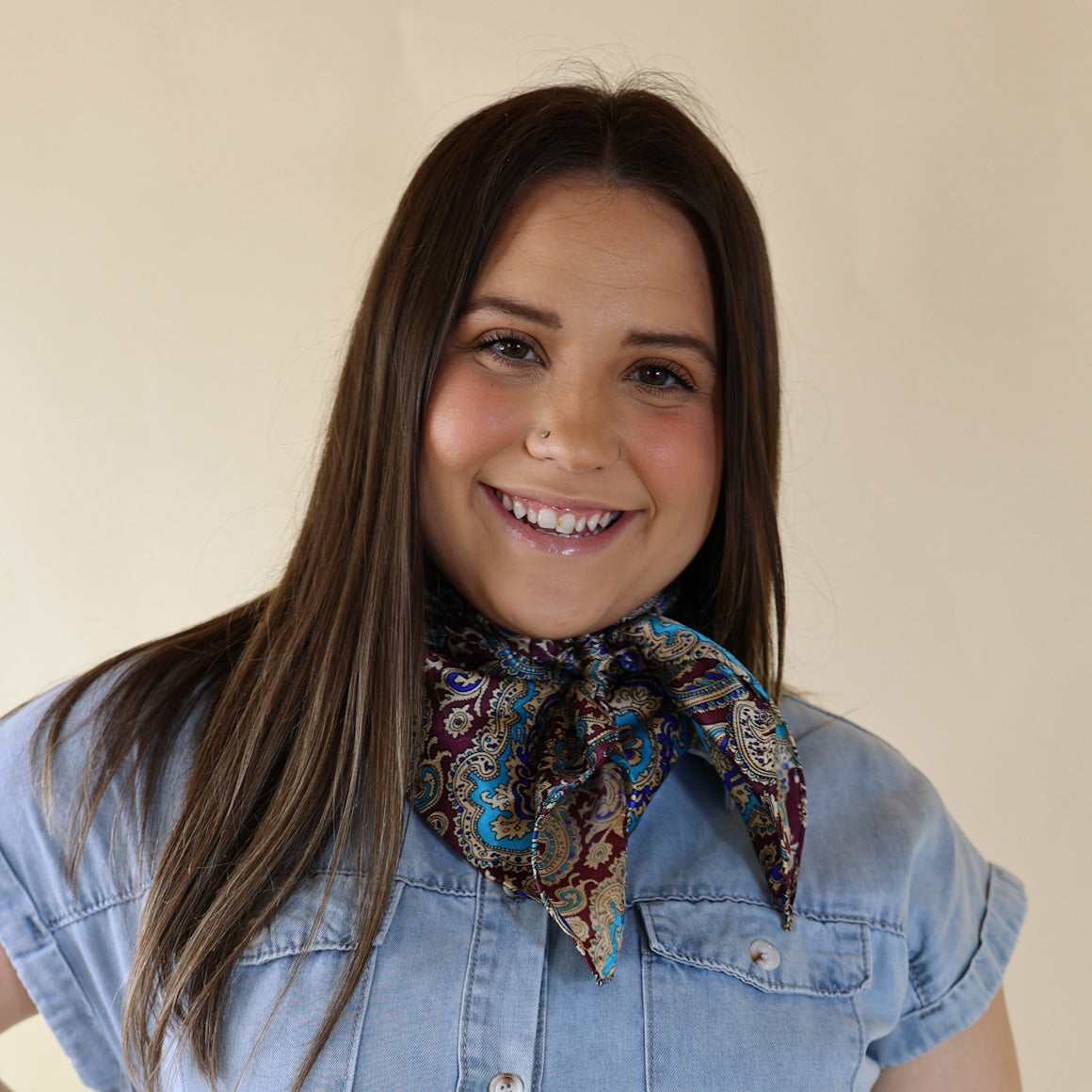 Brunette model wearing a short sleeve, denim button up with a red and gold paisly print scarf tied around her neck. This model is pictured in front of a beige background. 