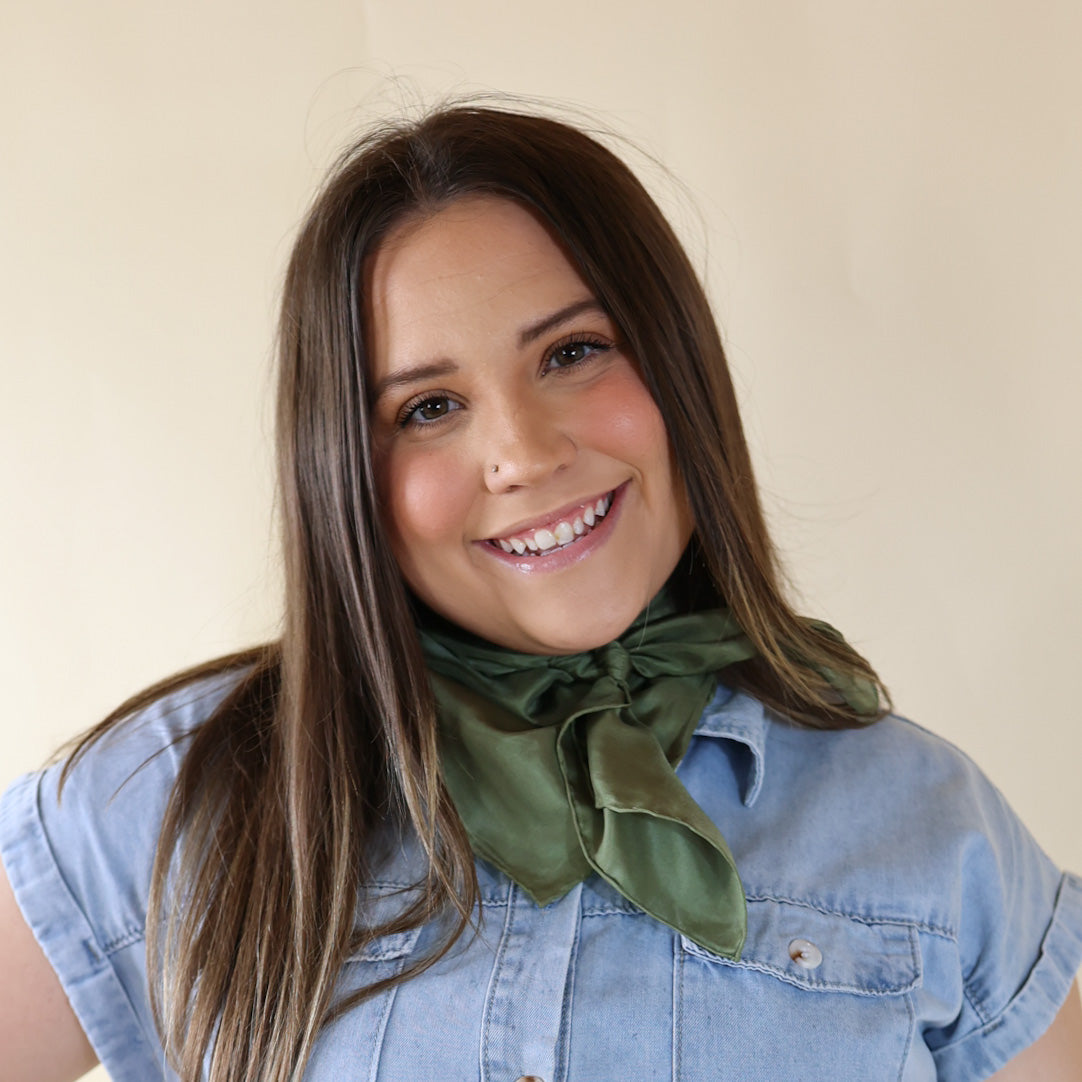 Brunette model wearing a short sleeve, denim button up with a sage green scarf tied around her neck. This model is pictured in front of a beige background. 