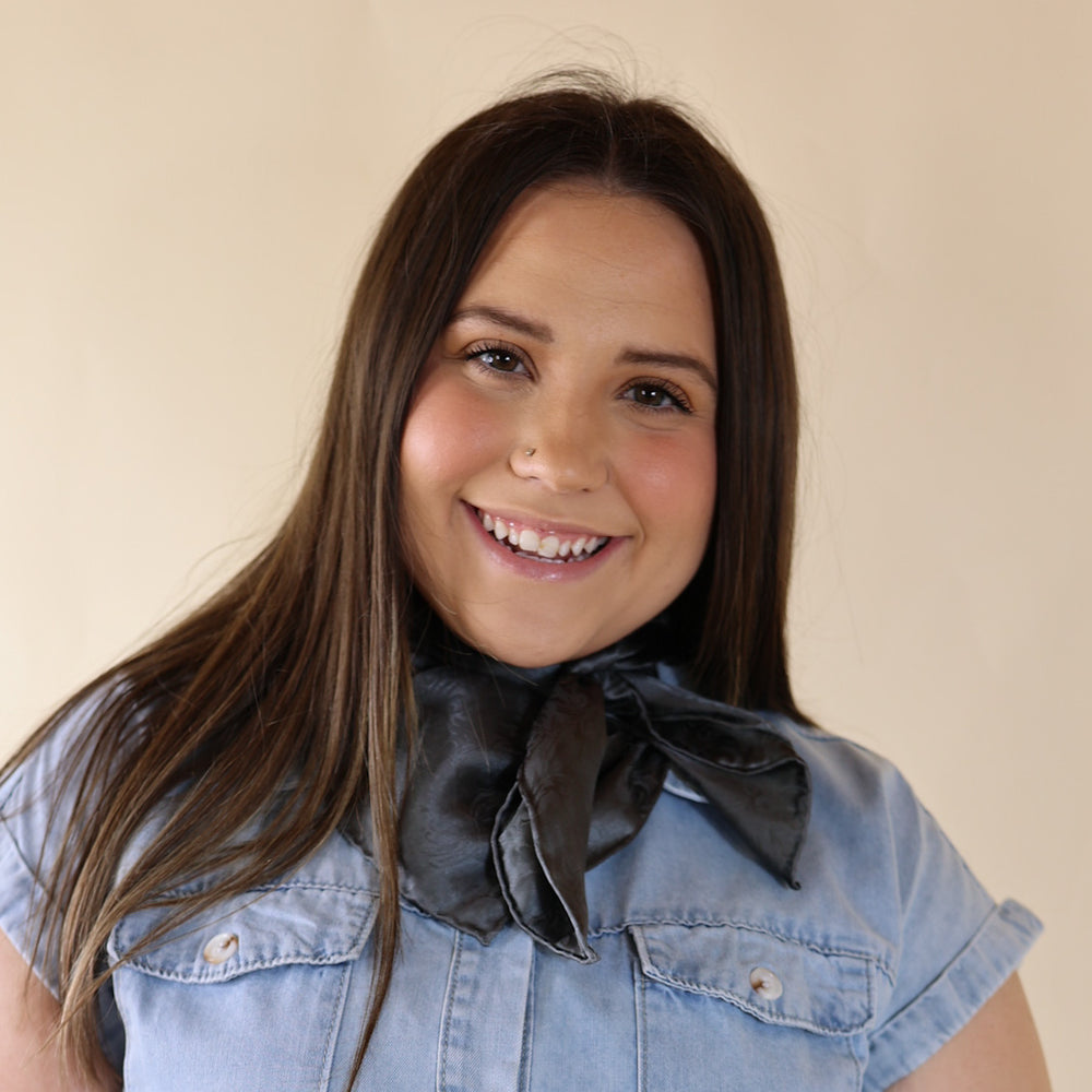 Brunette model wearing a blue dress with Jacquard Charcoal print scarf tied around her neck. Model is pictured in front of a beige background.