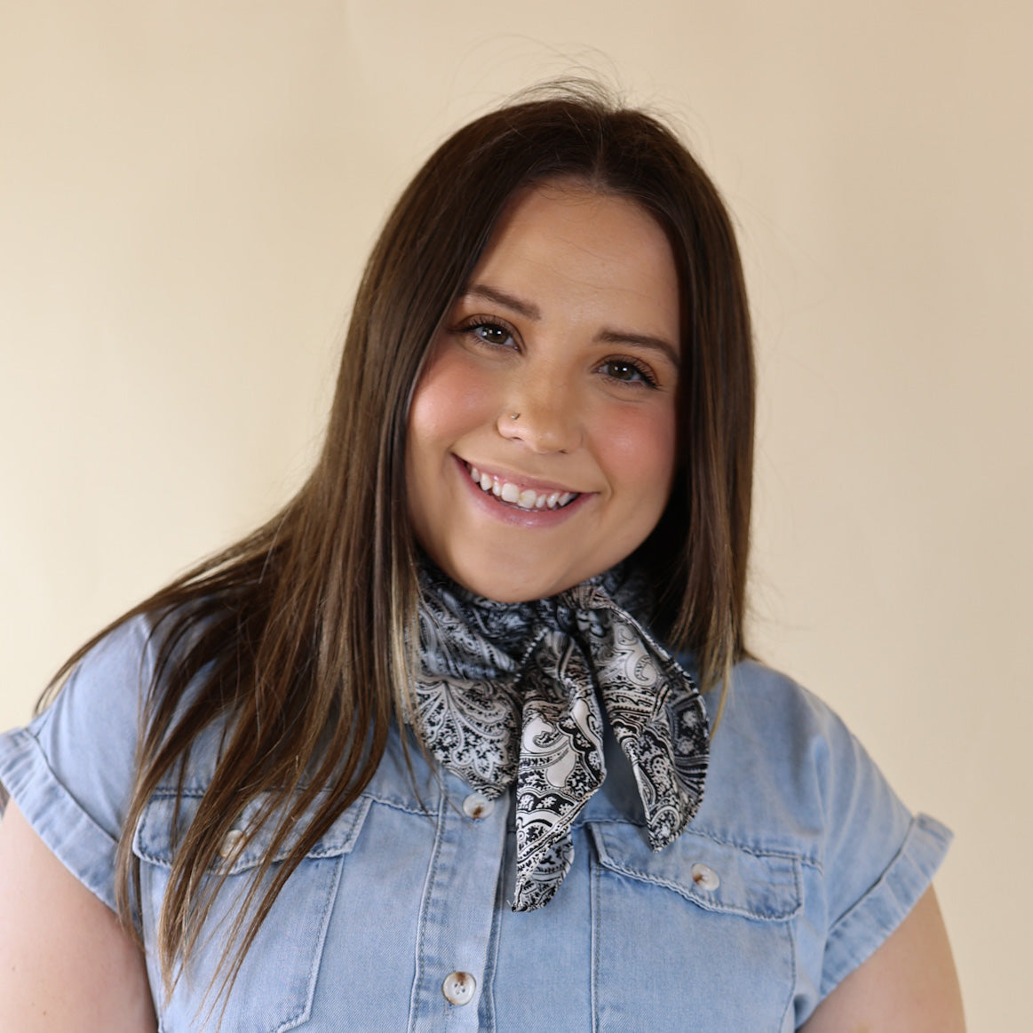 Brunette model wearing a short sleeve, denim button up with a black and white paisly print scarf tied around her neck. This model is pictured in front of a beige background. 