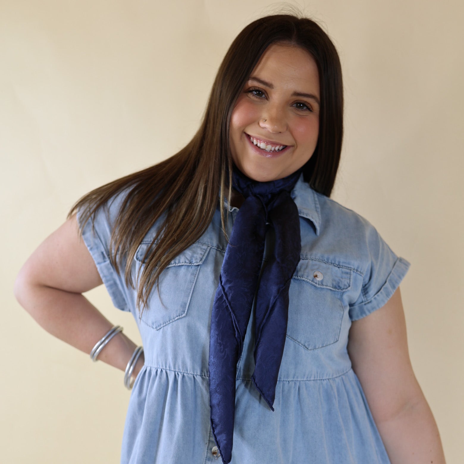 Brunette model wearing a short sleeve, denim button up with a navy jacquard print scarf tied around her neck. This model is pictured in front of a beige background. 