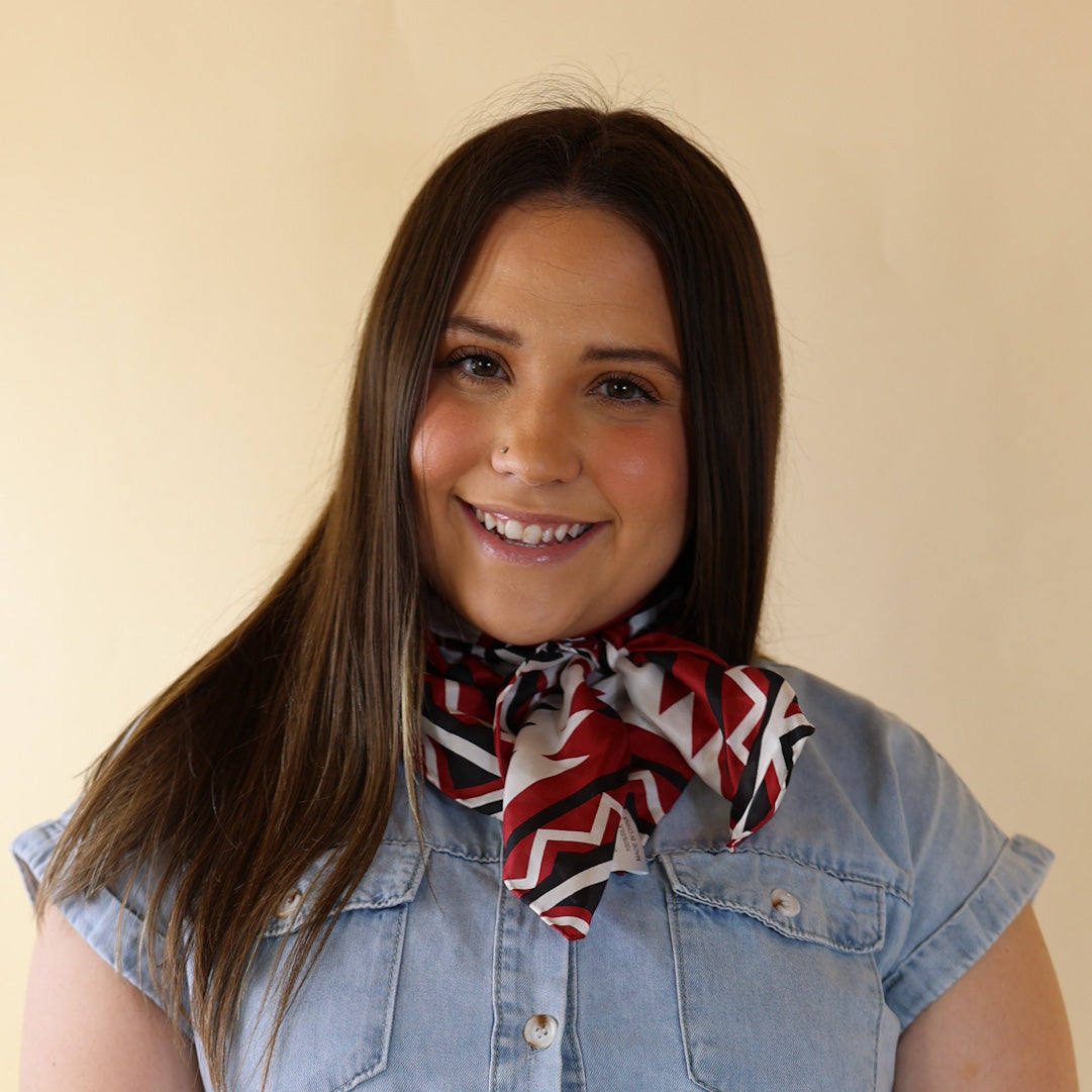 Brunette model wearing a short sleeve, denim button up with a black, white, and red aztec print scarf tied around her neck. This model is pictured in front of a beige background. 