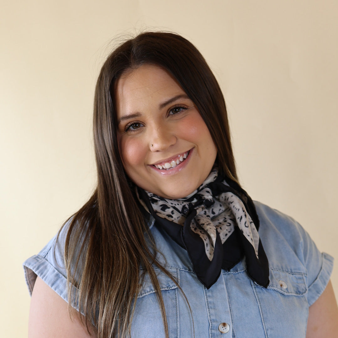 Brunette model is pictured wearing a denim button up top and a black and white scarf tied around her neck. She is pictured in front of a beige background. 