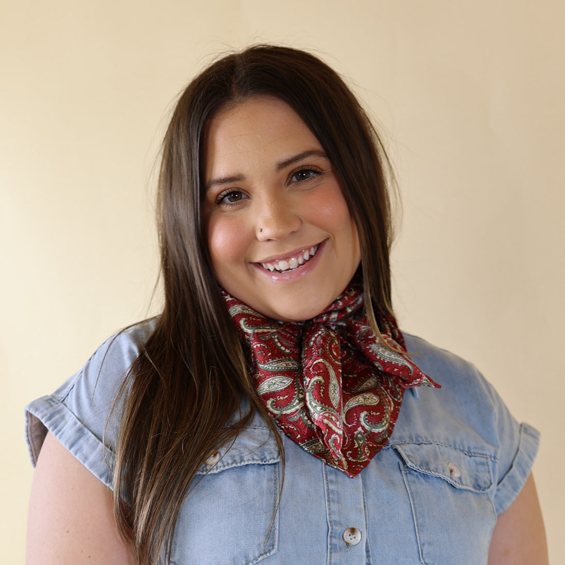 Brunette model wearing a short sleeve, denim button up with a wine and olive paisley print scarf tied around her neck. This model is pictured in front of a beige background. 
