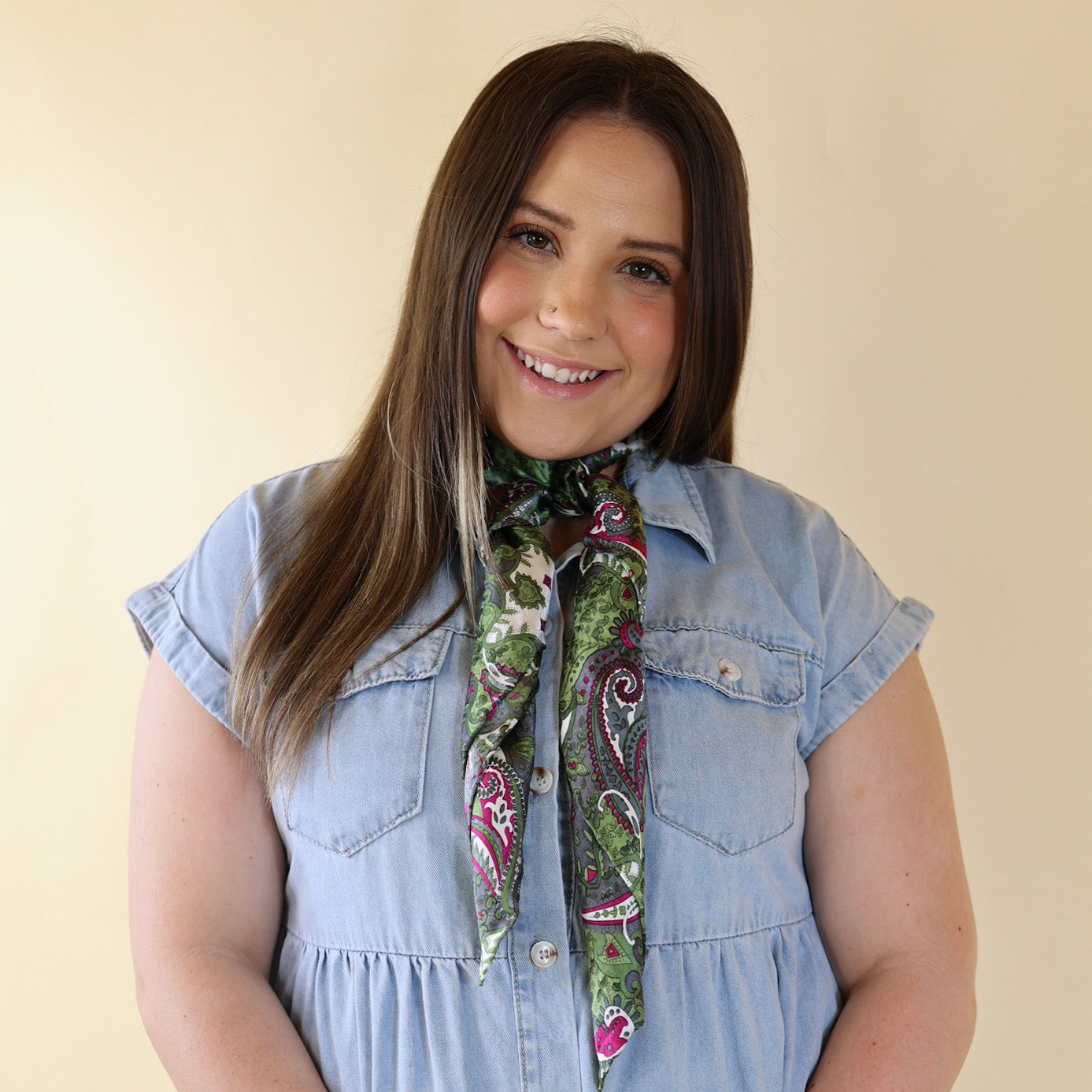 Brunette model is pictured wearing a denim button up top and a green, white, and pink printed scarf tied around her neck. She is pictured in front of a beige background. 