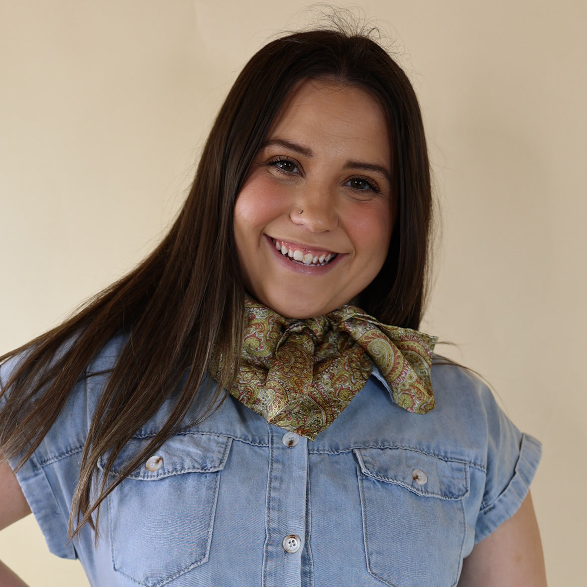 Brunette model is pictured wearing a denim button up top and a brass and brown paisley printed scarf tied around her neck. She is pictured in front of a beige background. 