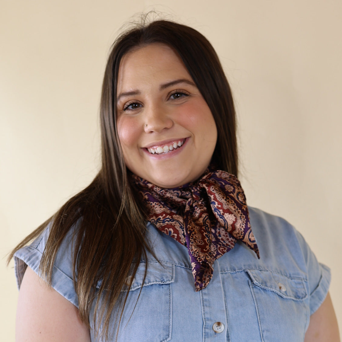 Brunette model wearing a short sleeve, denim button up with a pum and maroon paisley print scarf tied around her neck. This model is pictured in front of a beige background. 