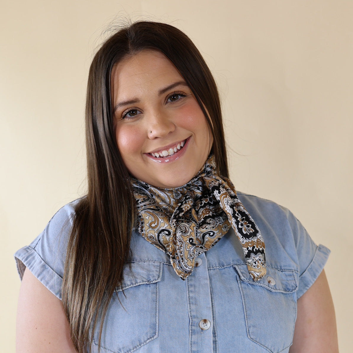 Brunette model is pictured wearing a denim button up top and a tan and black mix paisley printed scarf tied around her neck. She is pictured in front of a beige background. 