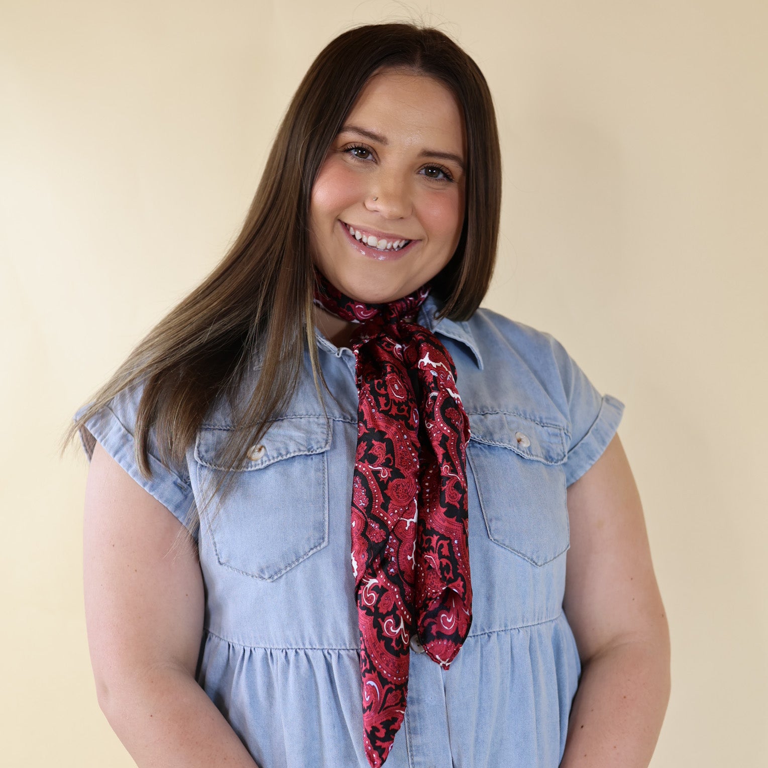 Brunette model wearing a blue dress with Red and Black Paisley print scarf tied around her neck. Model is pictured in front of a beige background.