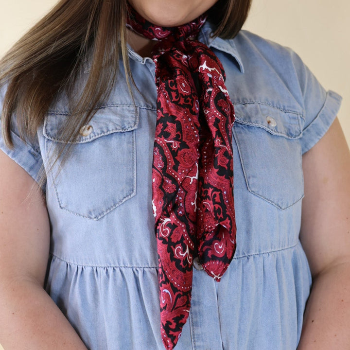 Brunette model wearing a blue dress with Red and Black Paisley print scarf tied around her neck. Model is pictured in front of a beige background.