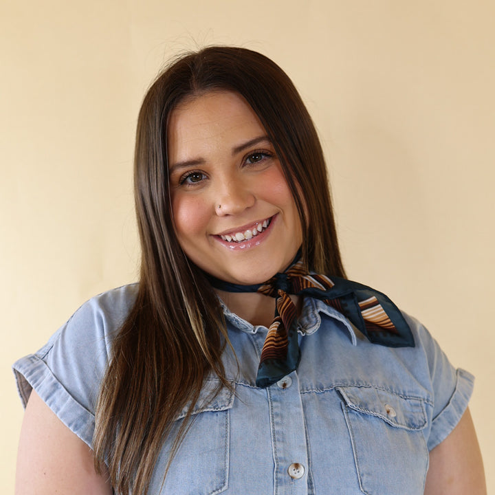 Brunette model is pictured wearing an ivory, leopard print long sleeve top and a blue and brown striped scarf tied around her neck. She is pictured in front of a beige background. 