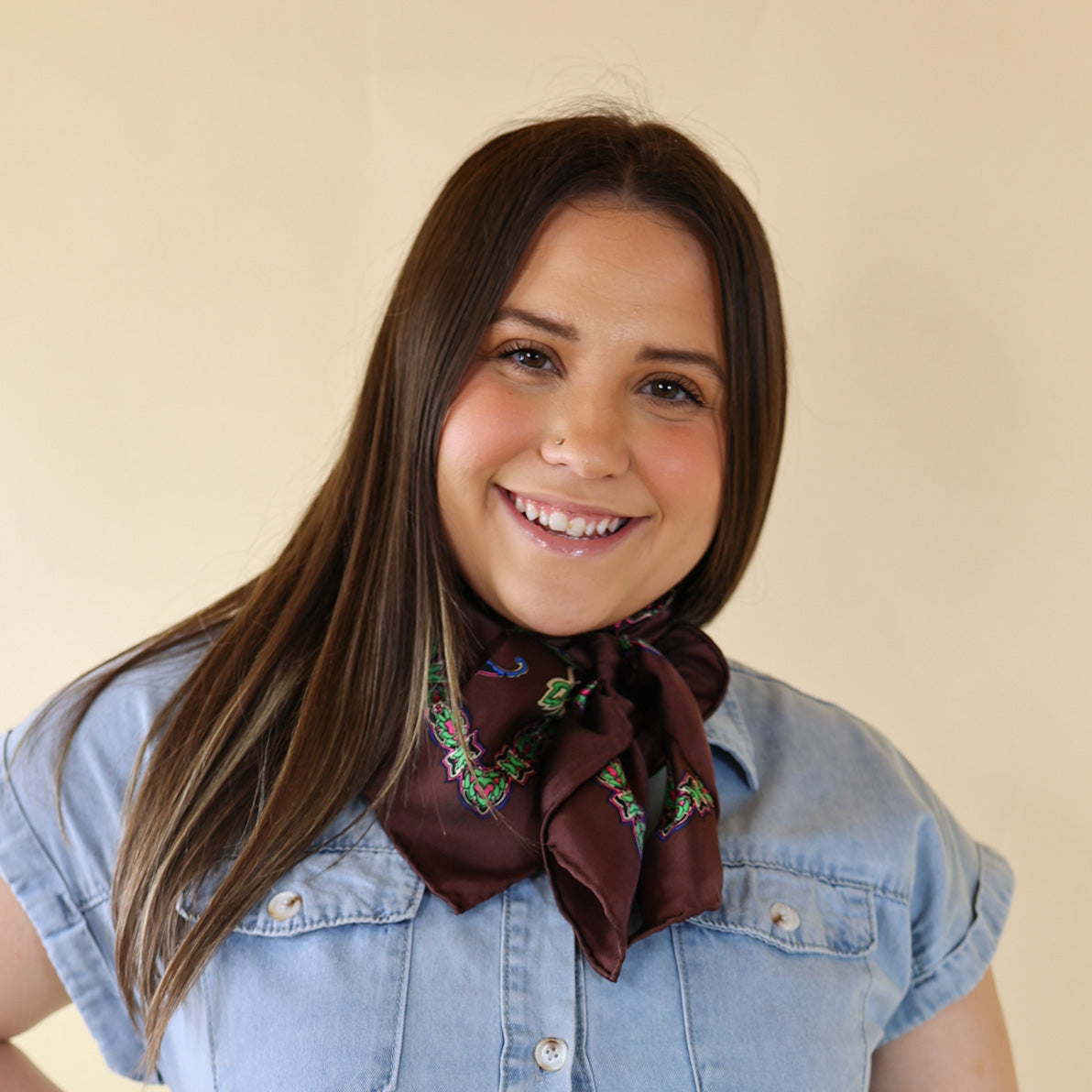 Brunette model is pictured wearing a denim button up top and a dark brown printed scarf tied around her neck. She is pictured in front of a beige background. 