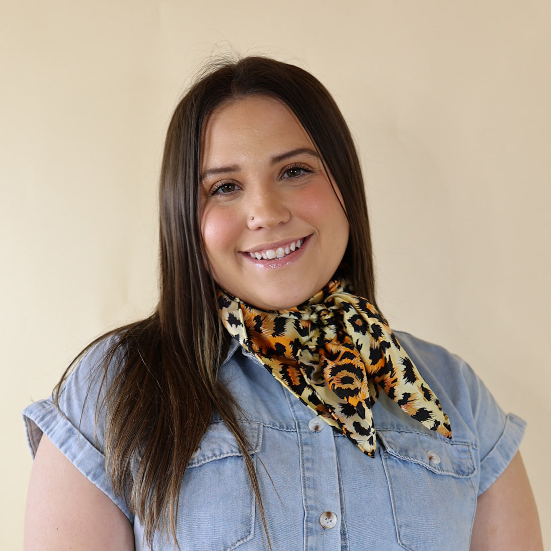 Brunette model wearing a short sleeve, denim button up with a cheetah print scarf tied around her neck. This model is pictured in front of a beige background. 