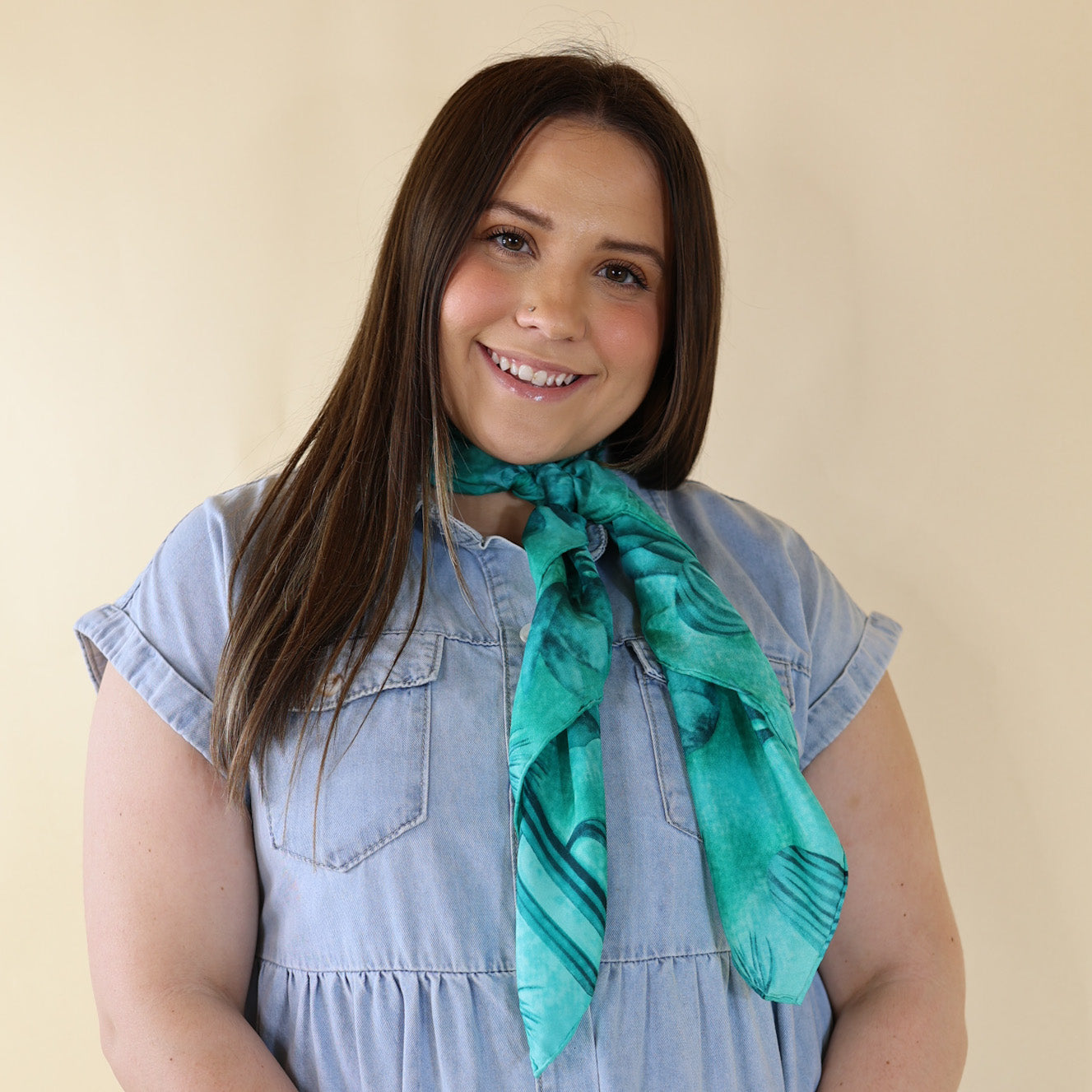 Brunette model is pictured wearing a denim button up top and a teal, cacti printed scarf tied around her neck. She is pictured in front of a beige background. 