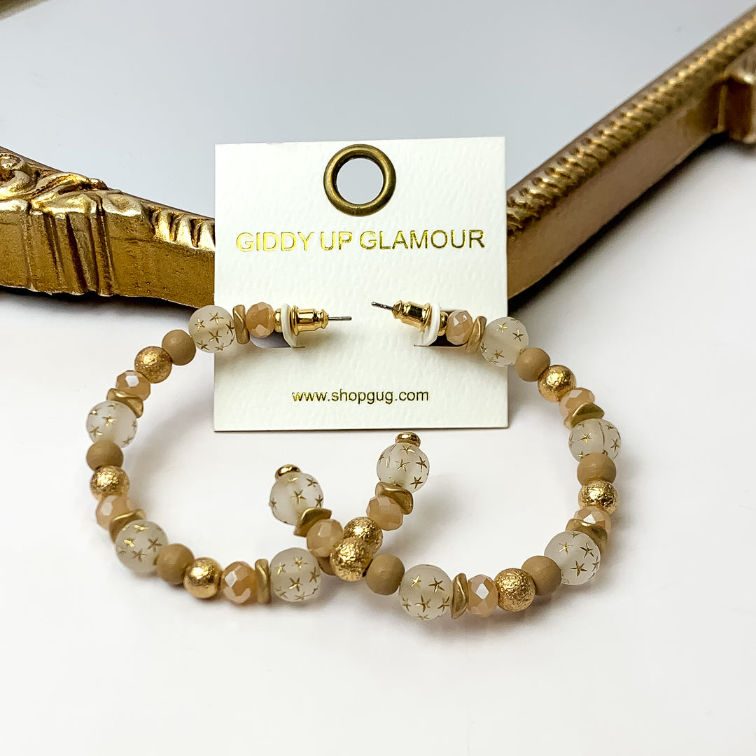 A pair of light brown and gold hoop earrings featuring gold stars on an ivory earring card. These earrings are pictured with a white background and gold mirror.