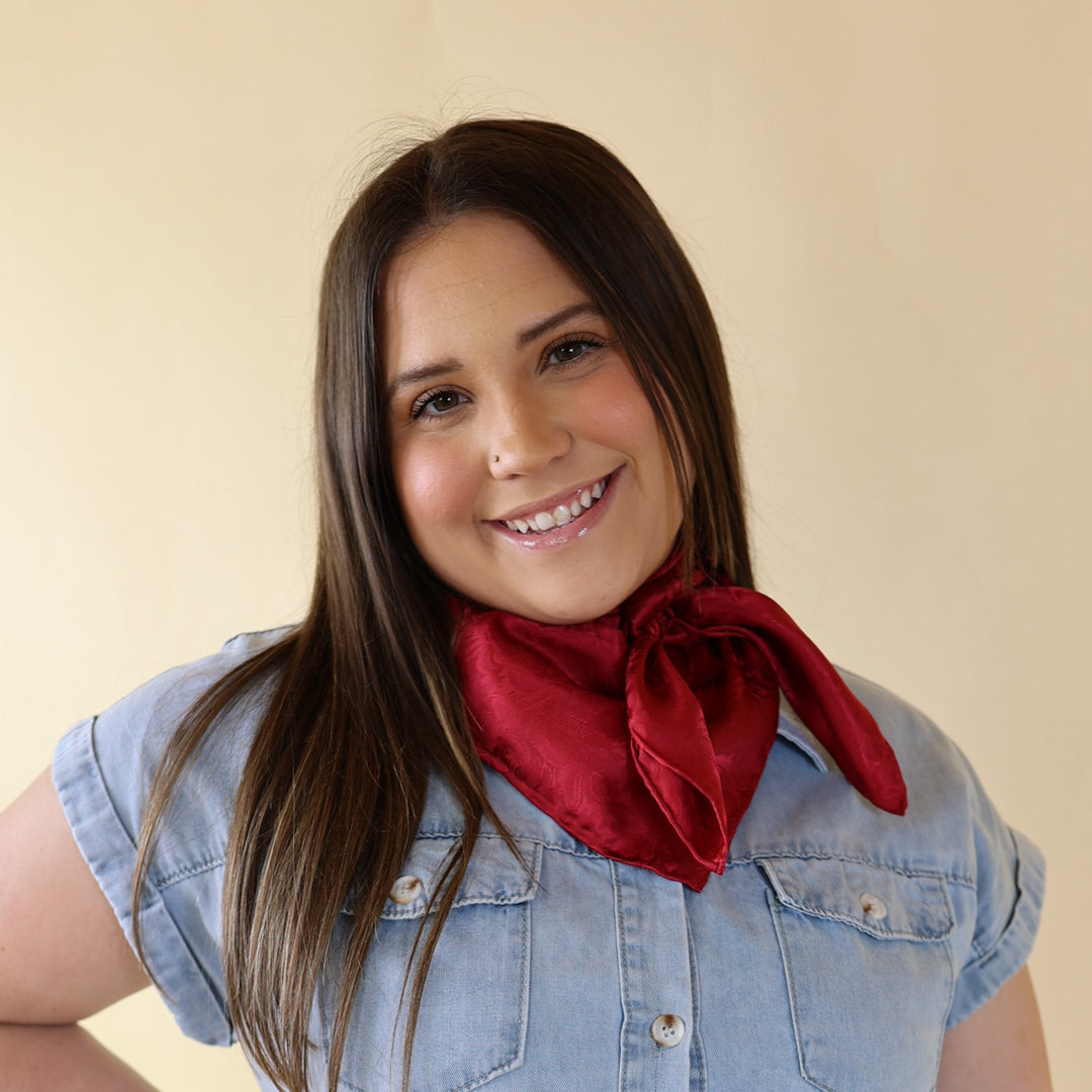Brunnet model is wearing a blue dress with a red scarf tied around her neck. Model is standing in front of beige background.