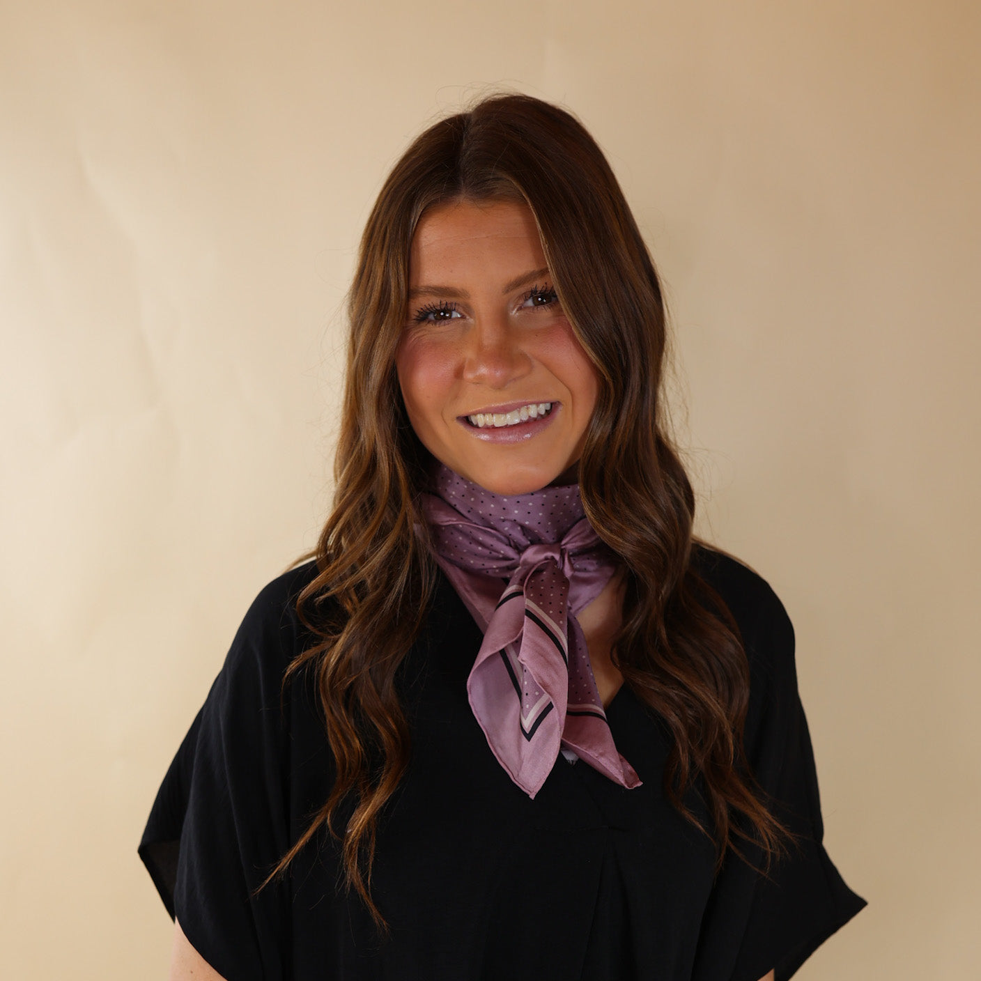 Brunette model is pictured wearing a Black, Drop shoulder top with a pink and Black Polka Dot scarf tied around her neck. Model is pictured in front of a beige background. 