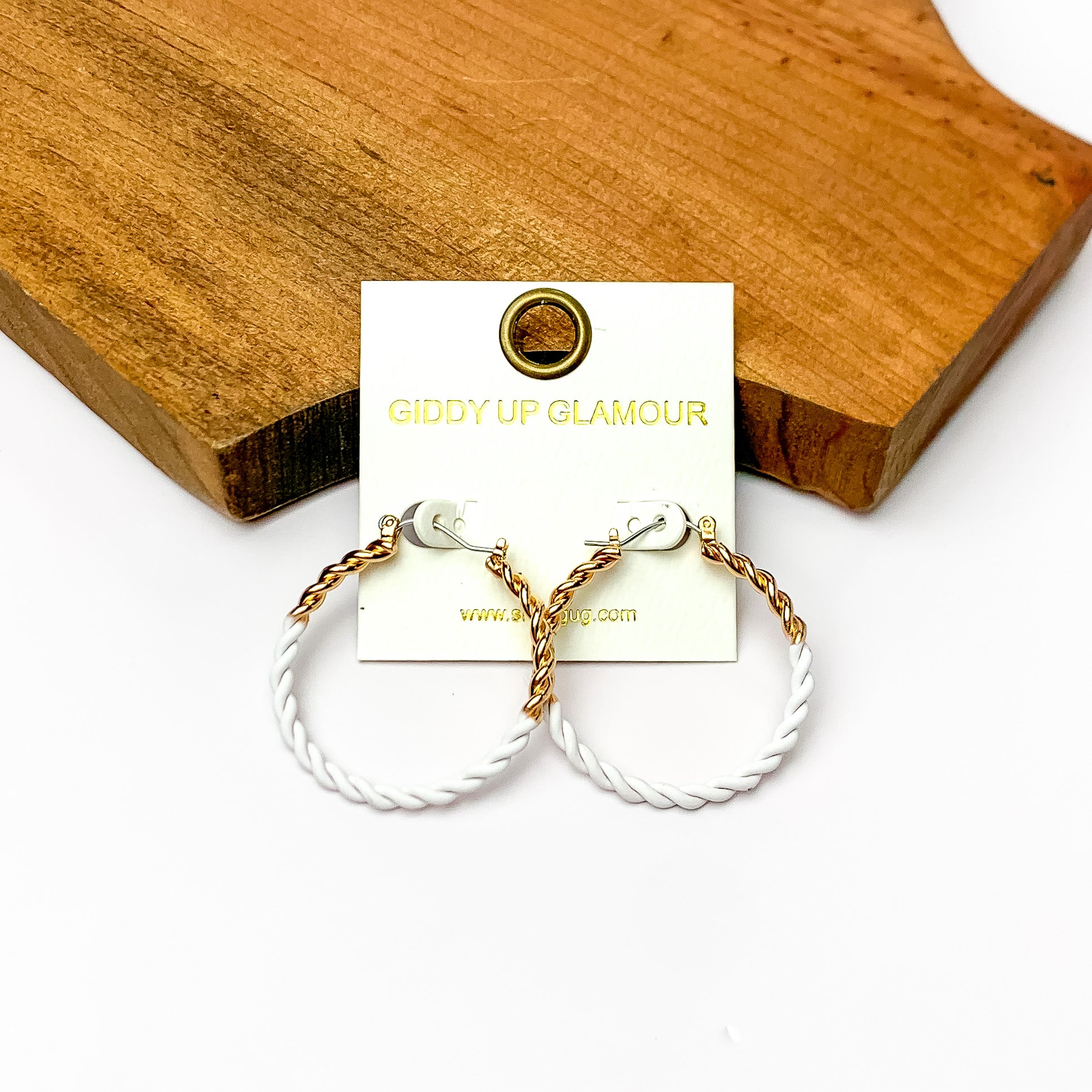 Twisted Gold Tone Hoop Earrings in White. Pictured on a white background with wood at the top.