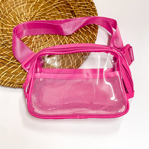 Pictured is a rectangle clear fanny pack with a fuchsia pink outline. This bag also includes a fuchsia pink strap, fuchsia pink accents, and fuchsia pink mesh pockets. This bag is pictured on a white and brown patterned background. 