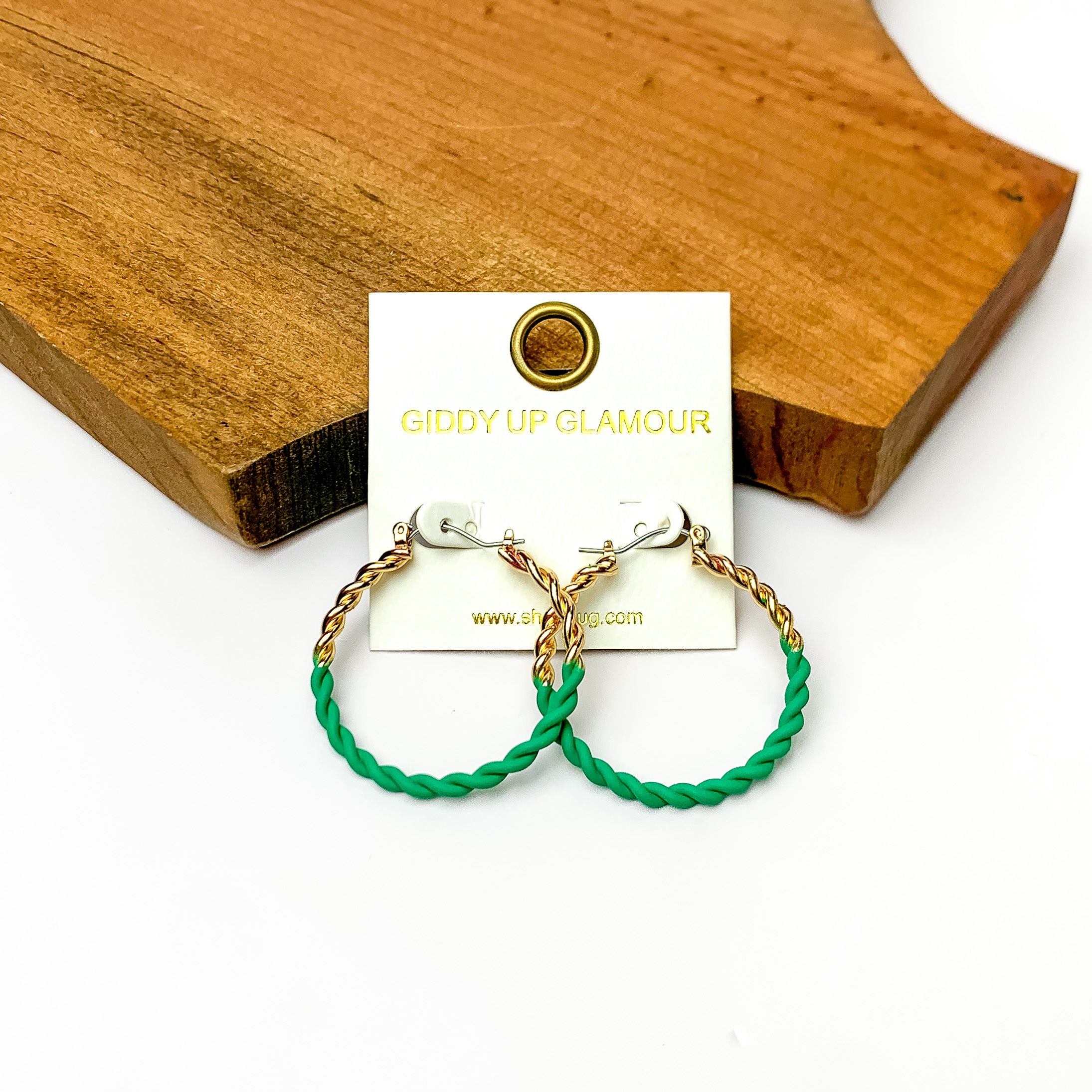 Twisted Gold Tone Hoop Earrings in Green. Pictured on a white background with a wood piece behind.