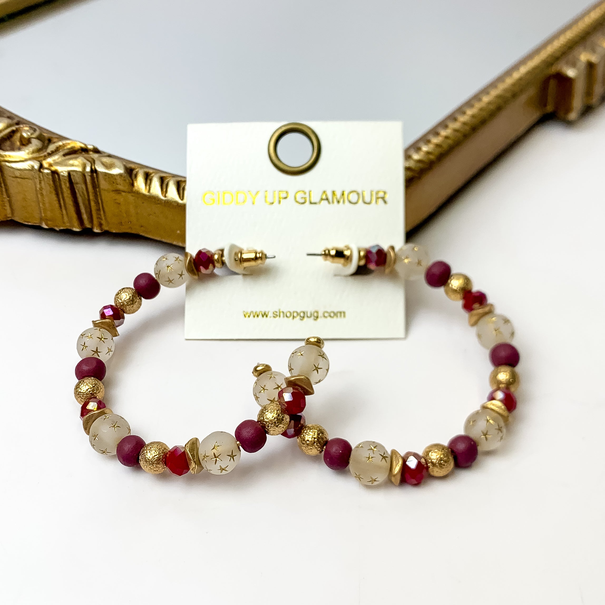 A pair of maroon and gold hoop earrings featuring gold stars on an ivory earring card. These earrings are pictured with a white background and gold mirror.