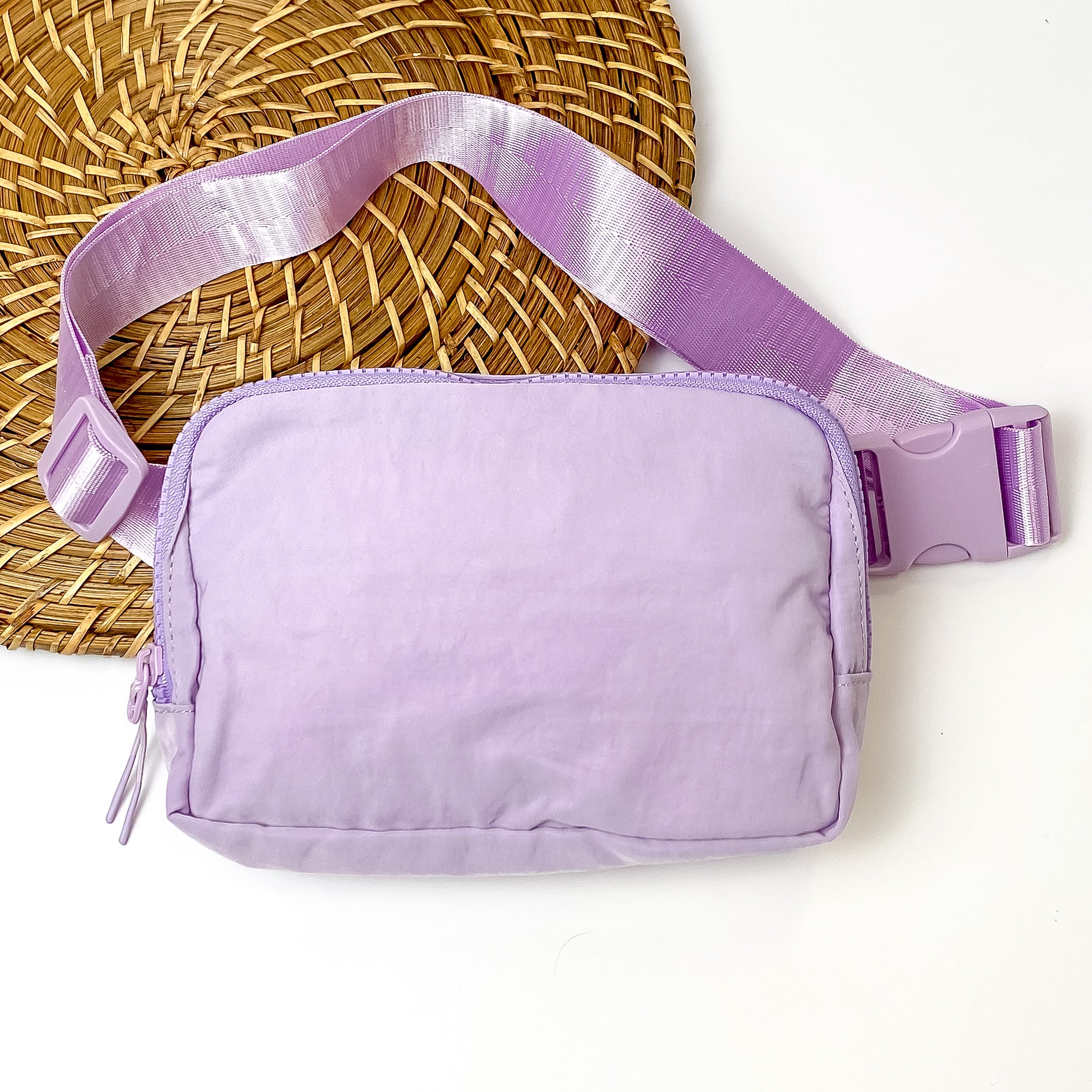 Pictured is a rectangle fanny pack with a top zipper with tassel in light purple. This bag also includes a light purple strap and light purple accents. This bag is pictured on a white and brown patterned background. 