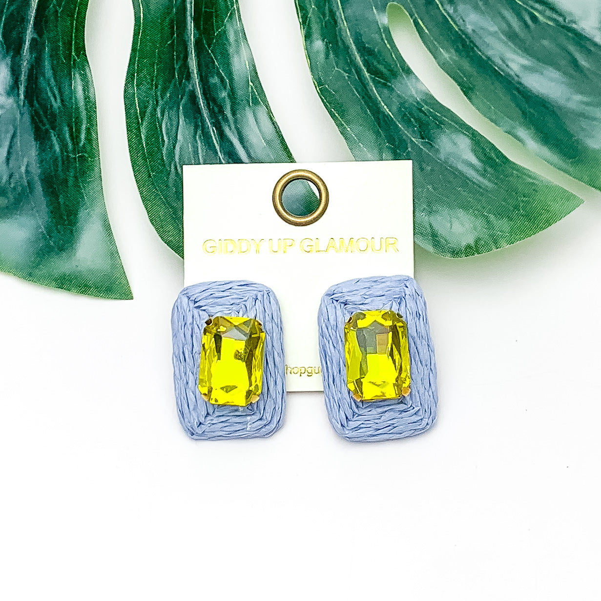 Truly Tropical Raffia Rectangle Earrings in Periwinkle Blue With Yellow Crystal. Pictured on a white background with the earrings laying on a large leaf.