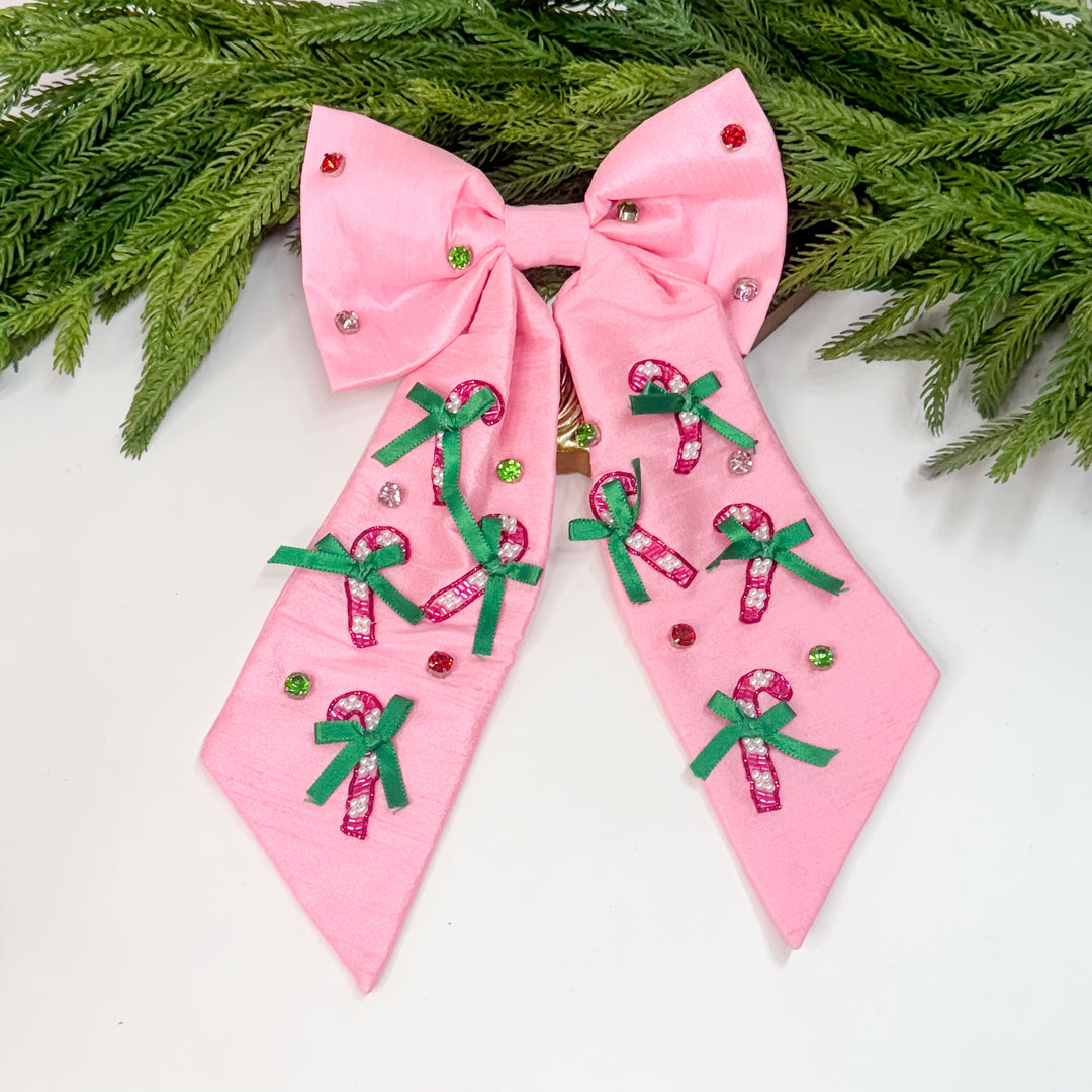 Oversized pink holiday hair bow with beaded candy canes, green ribbon bows, and gem embellishments displayed on greenery background.