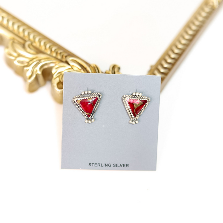 Triangular earrings with red gemstones on a gray card, gold necklace in the background