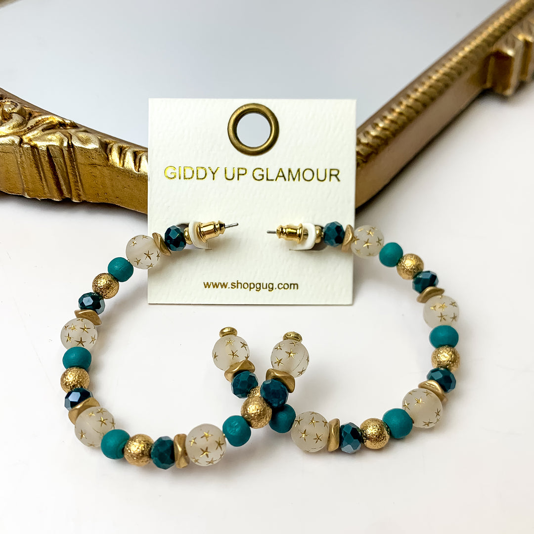 A pair of teal and gold hoop earrings featuring gold stars on an ivory earring card. These earrings are pictured with a white background and gold mirror.
