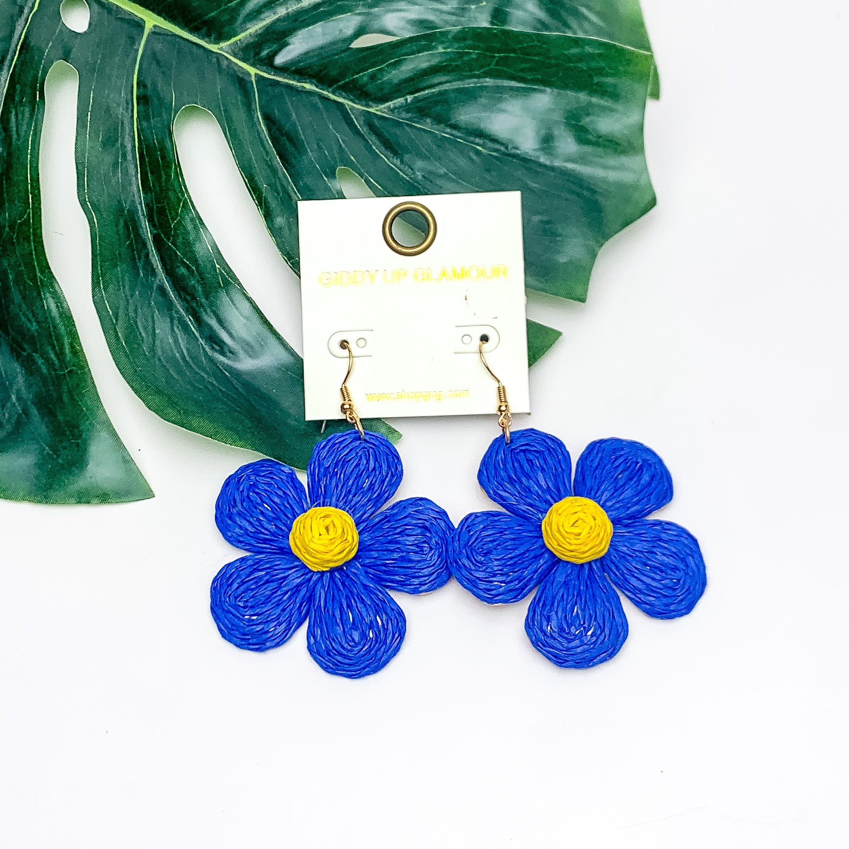 Darling Daisy Raffia Wrapped Flower Earrings in Blue. Pictured on a white background with the earrings laying on a large leaf.