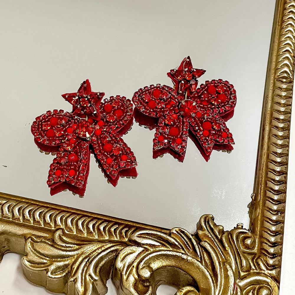 Red bow-shaped earrings with gold accents in front of a gold frame.