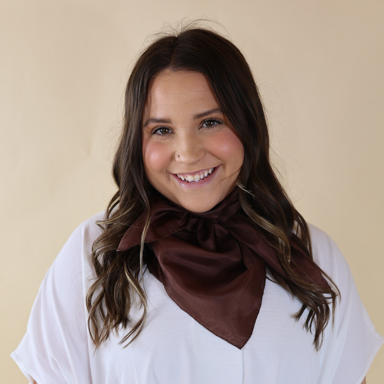 Brunette model is pictured wearing a white, Drop shoulder top with a solid brown scarf tied around her neck. Model is pictured in front of a beige background.