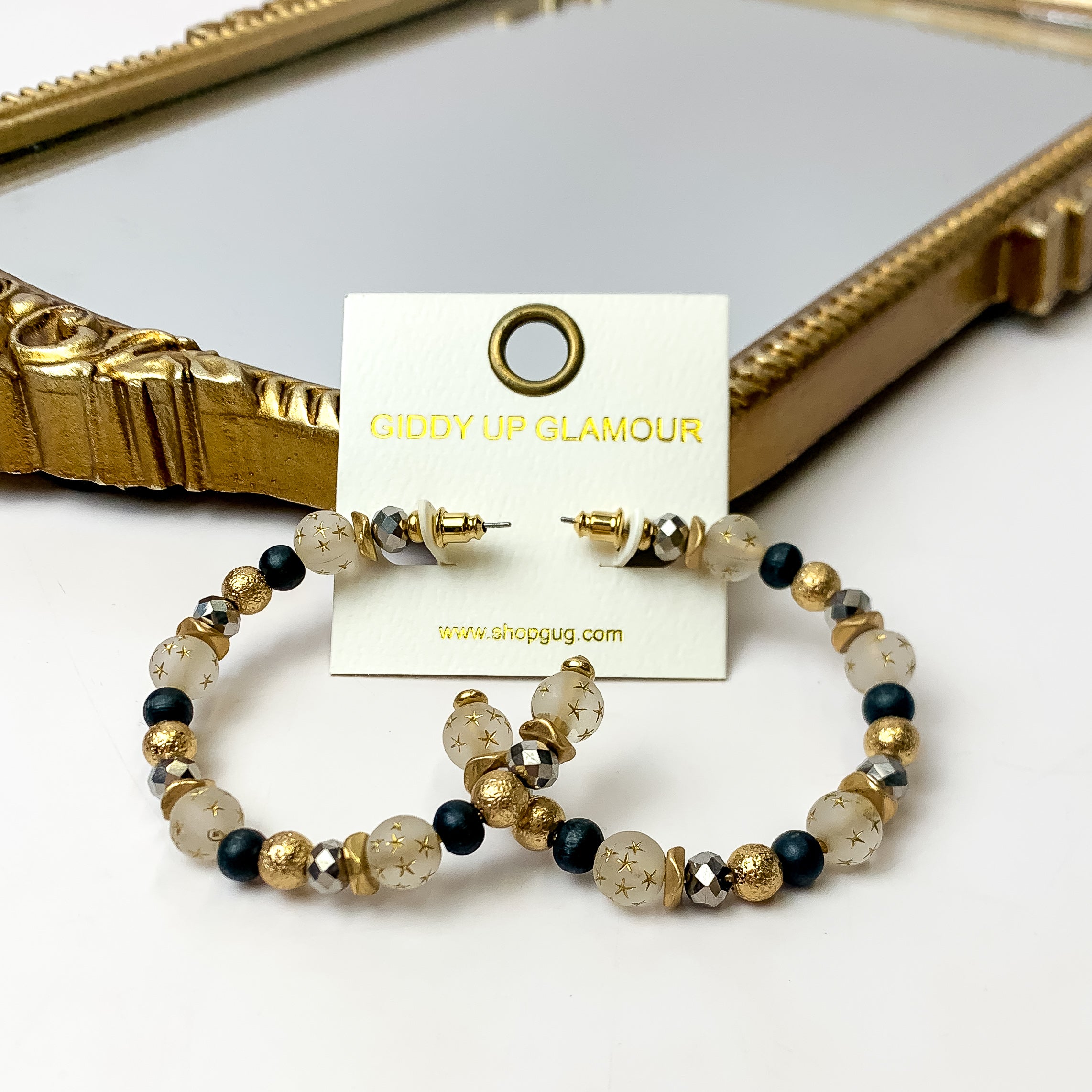 A pair of black and gold hoop earrings featuring gold stars on an ivory earring card. These earrings are pictured with a white background and gold mirror.