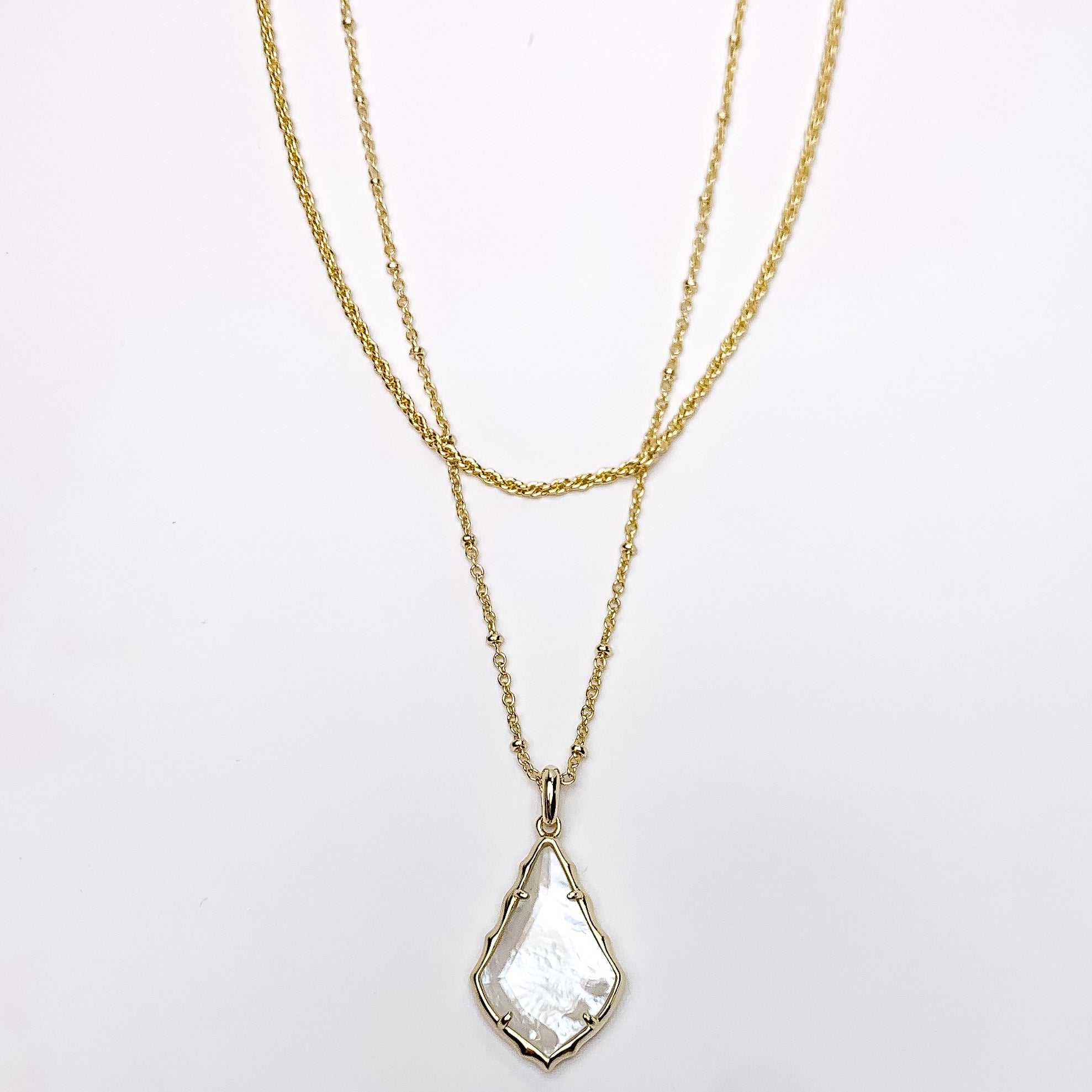 Two gold chain necklaces with the longer strand that has a gold and ivory mother of pearl pendant. This necklace is pictured on a white background. 